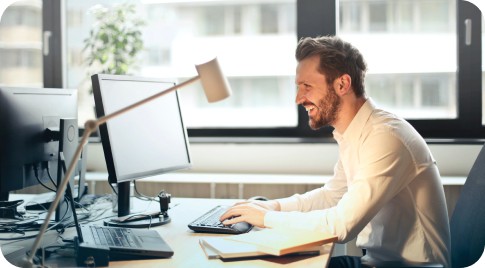 This is an image of a business man in a dress shirt sitting at the computer while smiling and working.
